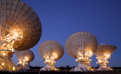The back of five large white radio telescope dishes, lit up at night