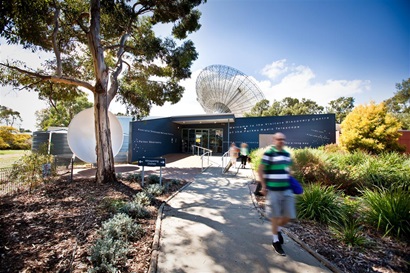 A person walking along a path in front of a blue building, a radio telescope dish is in the background.