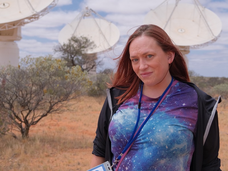 Woman in multi-coloured galaxy shirt smiles at the camera. Three radio telescope dishes are behind her.