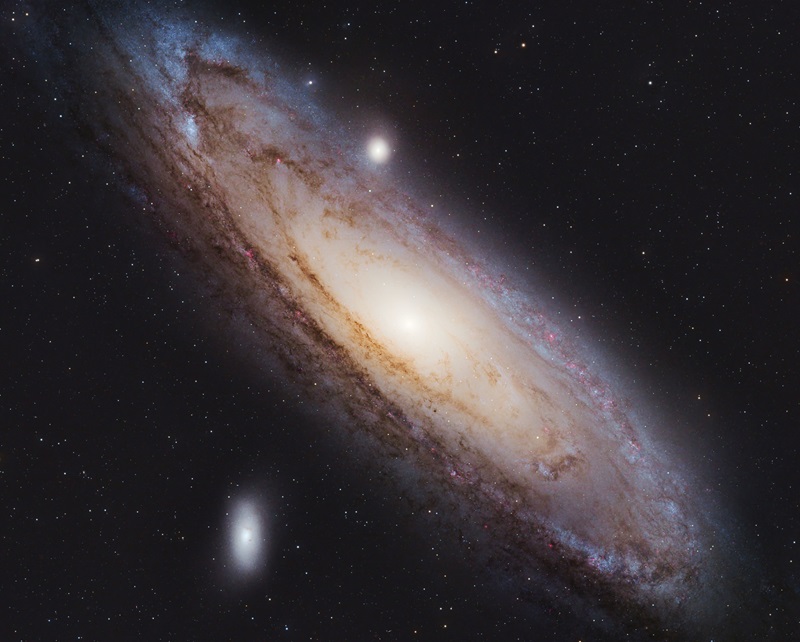 A background of stars. Large spiral of gas, dust and stars can be seen at roughly 45-degree angle stretching diagonally across the image from top left to bottom right. With two much smaller ovals of stars on the top and bottom edges of the larger galaxy roughly in the middle of the image. 