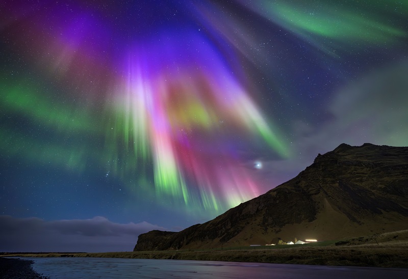 Curtains of rainbow colours hang down from the top of the image. Water and a mountain are seen in the foreground at the bottom of the image and the lights of a village can be seen at the base of the mountain. 