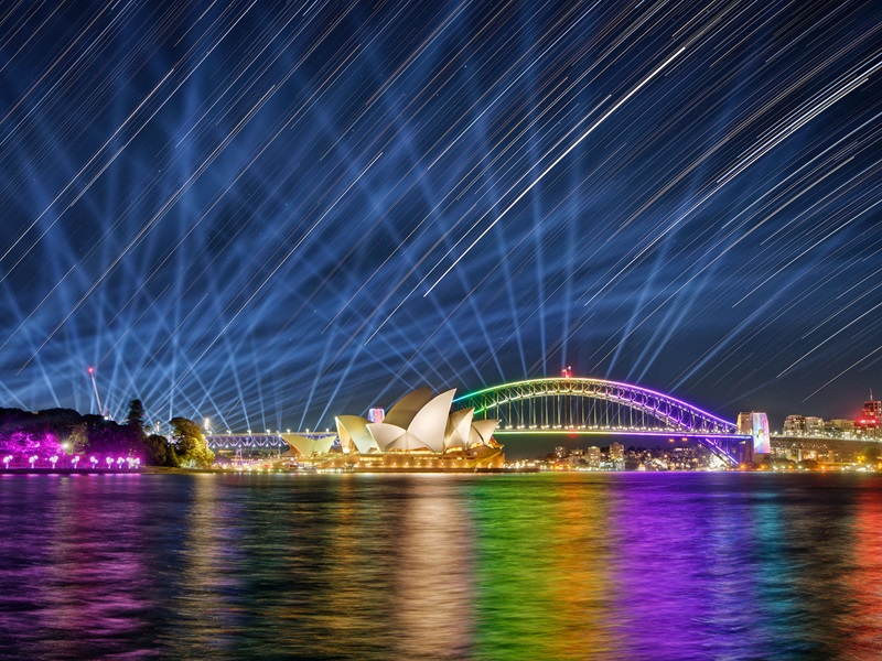 The top two thirds of the image shows streaks of light, a combination of the lights from the vivid light show and star trails. The bottom third of the image shows the Sydney Harbour Bridge, Opera House and harbour lit up in rainbow colours. 