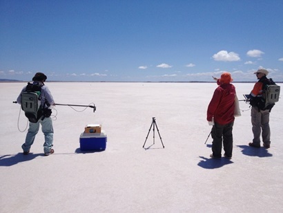 Three researchers stand on a vast desert plain at the Pinnacles, WA. At least a scientist holds a measurement tool above a white square used for calibration, with a tripod at centre. Two researchers look off into the distance at the right of the image.