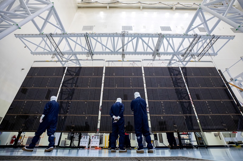 three people in blue overalls and hair nets stand in front of five columns of solar panels hanging vertically from a gantry in a large warehouse. 