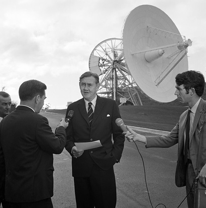 A black and white photograph of a man in a suit facing the camera, he's holding a piece of paper; three other men are holding mircophones and facing towards their interviewee; a large antenna dish is in the background.