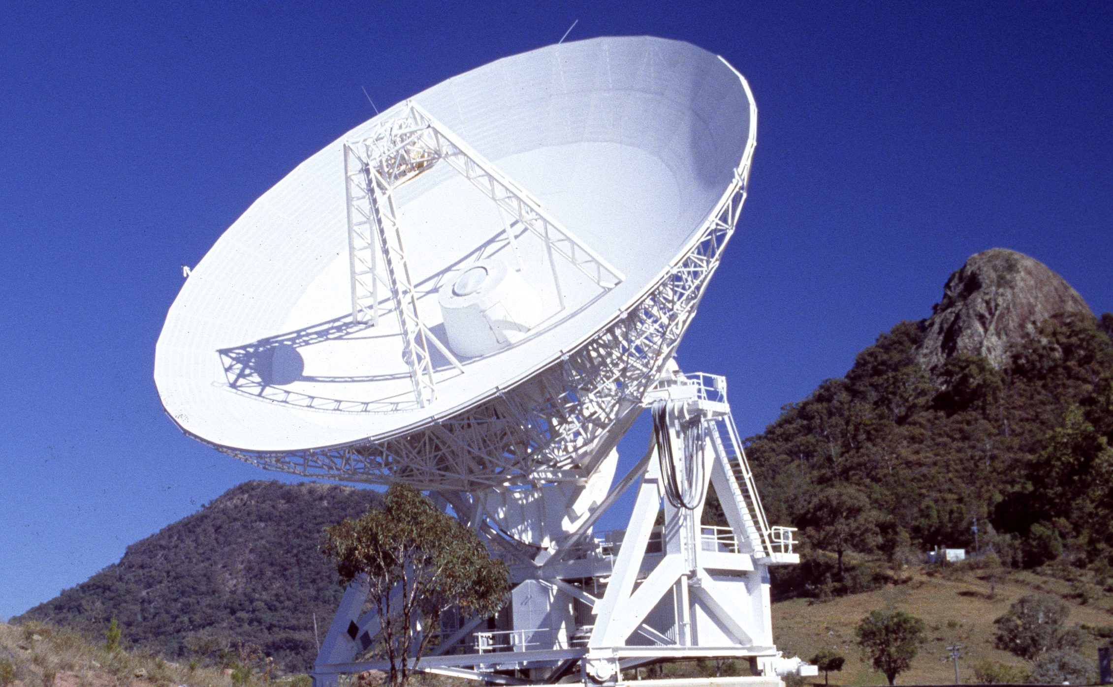 Image shows the Mopra radio telescope. It is a white radio astronomy telescope with a large rocky outcrop and blue sky in the background.