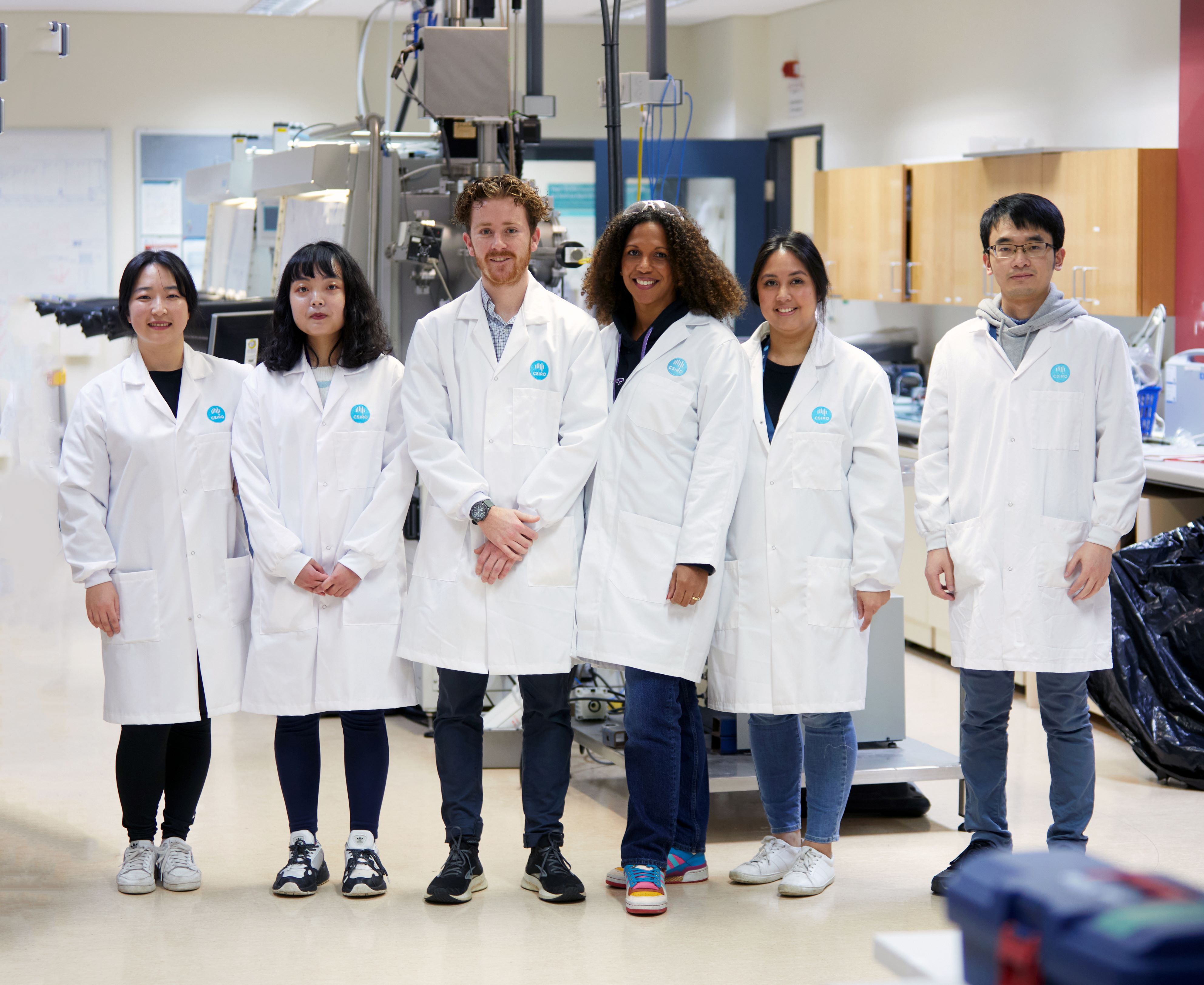 Group photo of four women and two men wearing lab coats in a lab, smiling directly at the camera