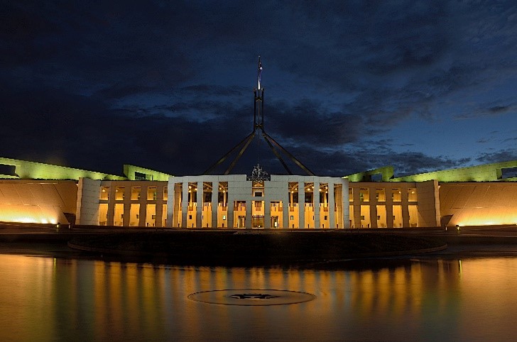 The image shows the Australian Parliament House in the evening. The shot looks out over water that has a single ripple in the centre. Parliament house is lit in a warm yellow light, while the light reflects of the roof in a lime green. The sky above is a dark blue with grey clouds.