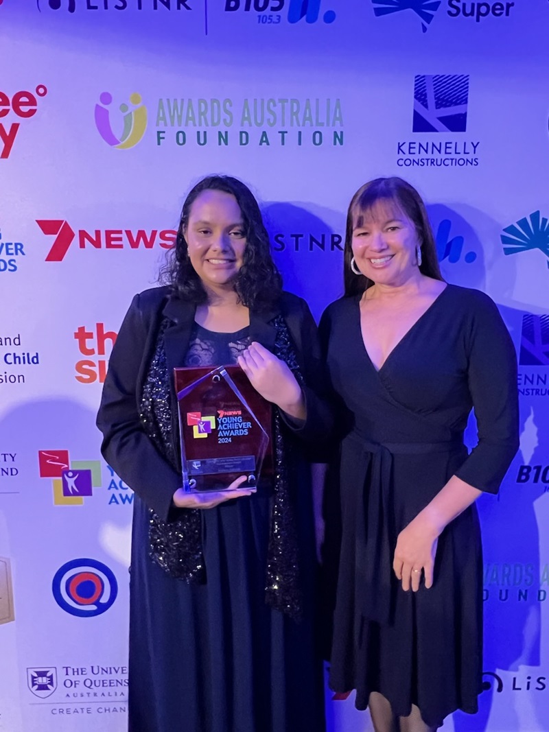 Two Indigenous women stand side by side smiling. The young woman is holding her award trophy.