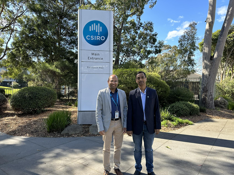 Two men standing outside in front of a large sign board 