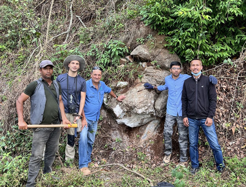 A group of people standing outdoors in front of rock and vegetation in the background, one person carries a pick axe