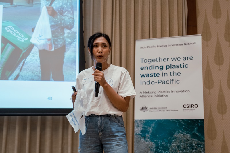 Woman holding microphone in front of a banner, presenting at a conference. 