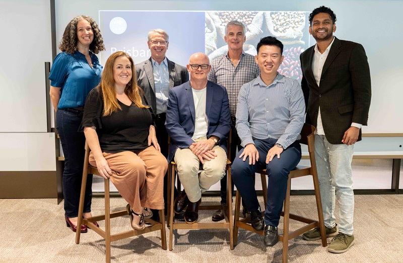 Group of people, three people seated in the front row and four people standing in the back row, smiling into the camera with a digital screen behind them