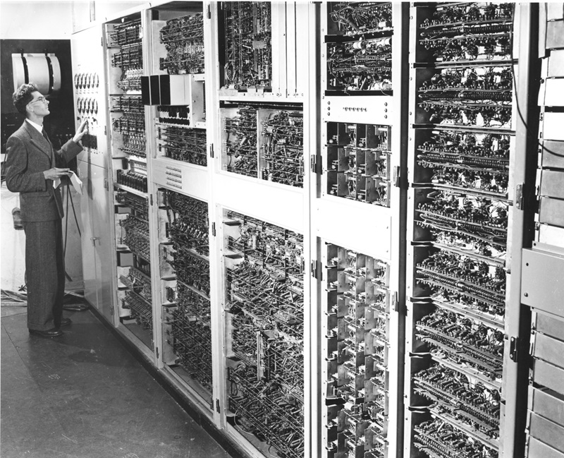 Black and white archive photograph of a man standing to the left next to CSIRAC a large machine covering an entire wall full of wires and cables 