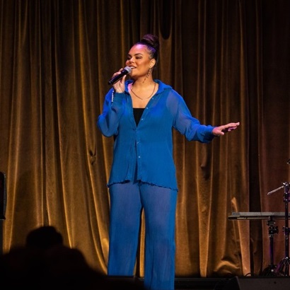 Mi-Kaisha performing on stage, holding a microphone and standing in front of golden curtains