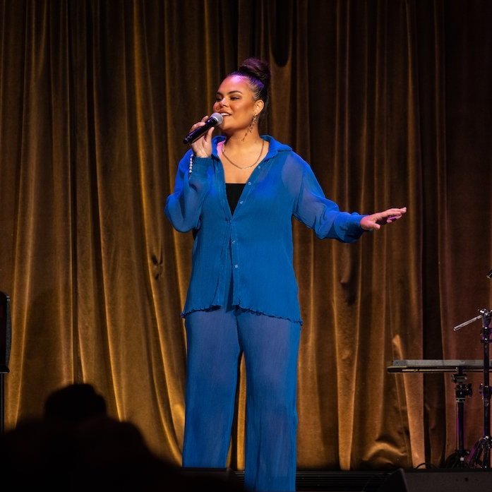 Mi-Kaisha performing on stage, holding a microphone and standing in front of golden curtains