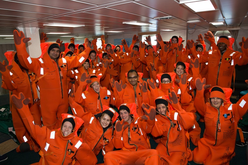 A large group of people in colourful immersion suits raise their arms into the air.