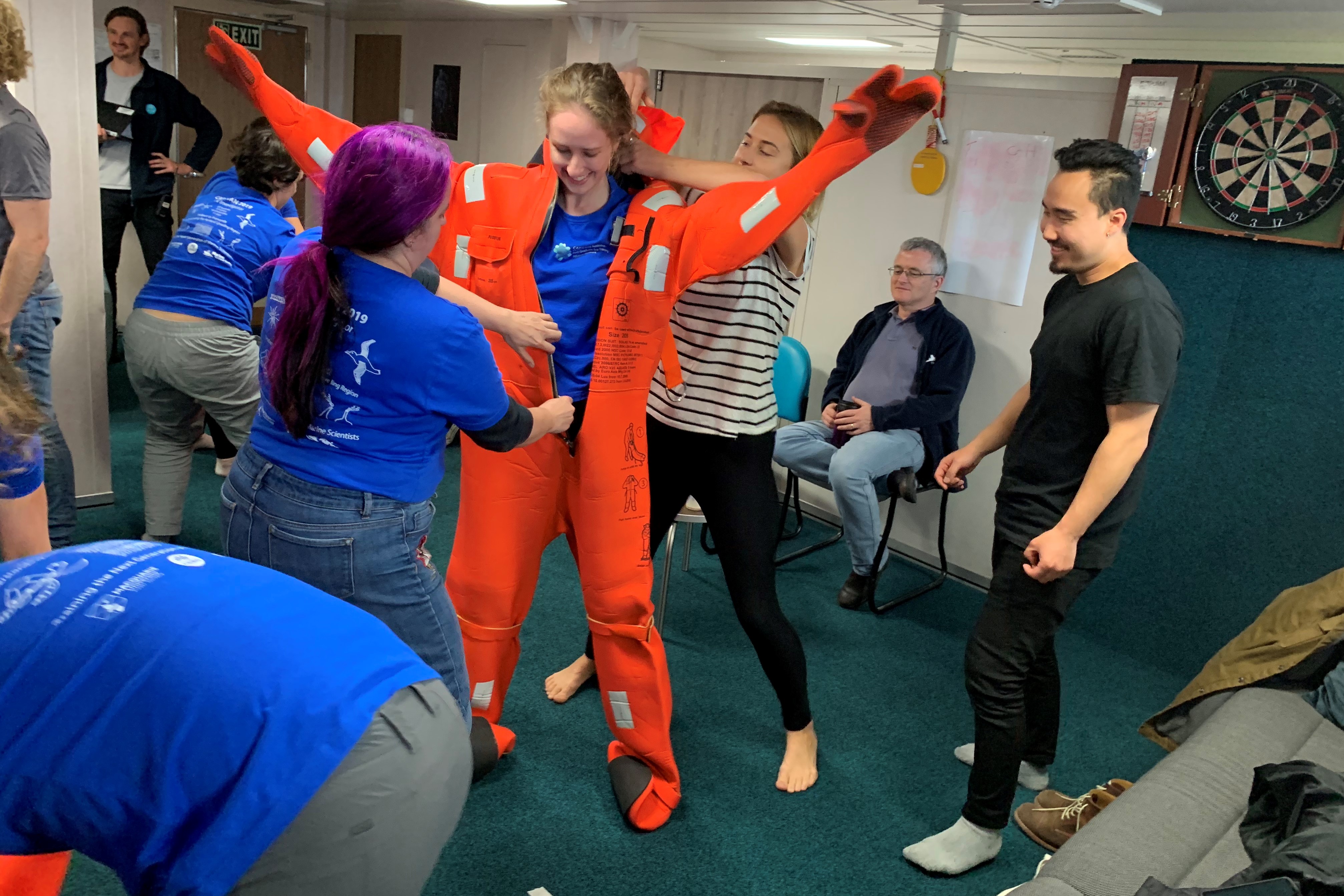 A person being helped to put on a bright orange safety suit by another person while other people stand around and watch.