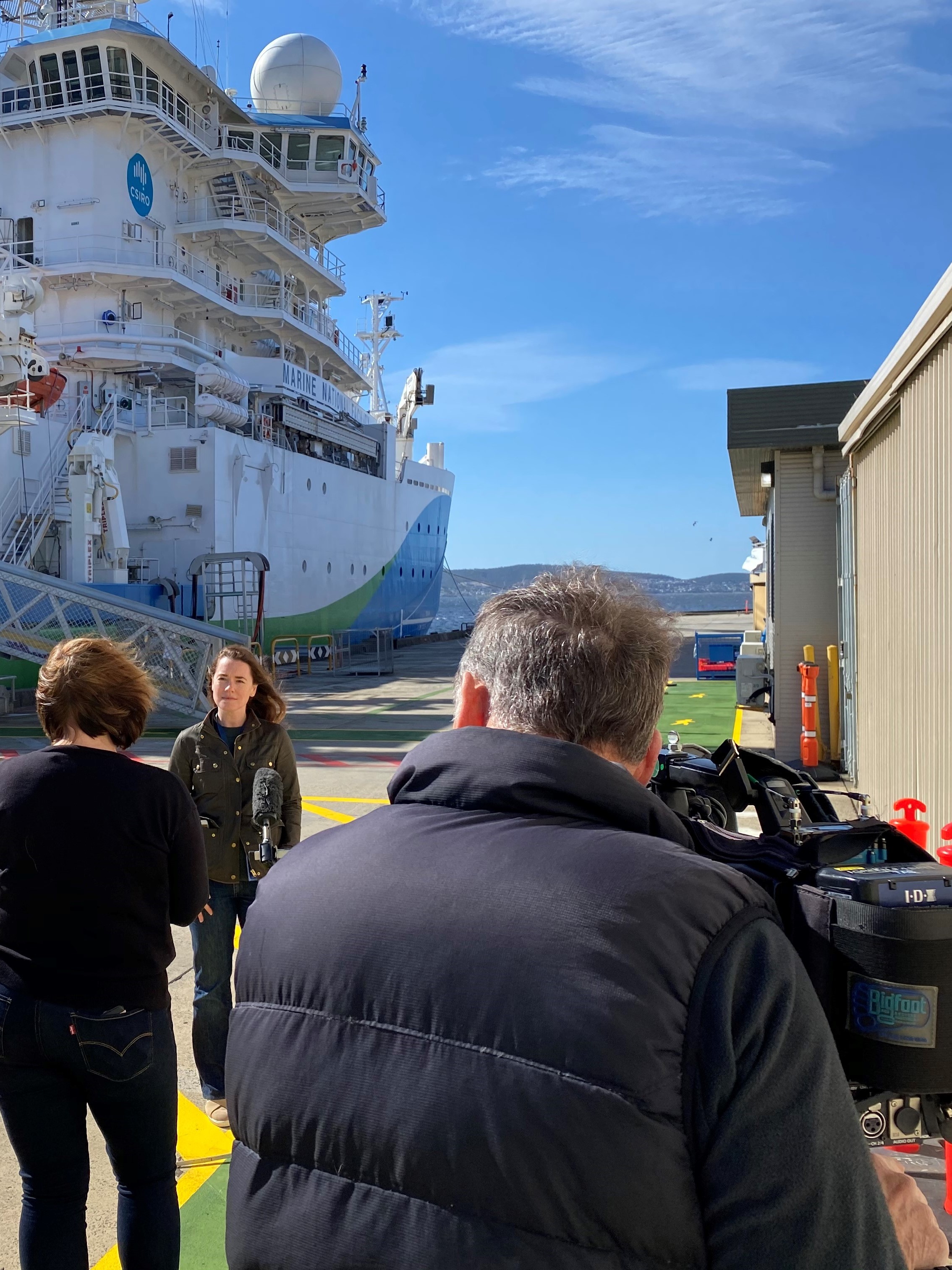 A person standing on a wharf next to a ship being interviewed by a reporter and cameraman.