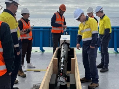 People standing around a piece of scientific equipment in a wooden box on the deck of a ship at sea.