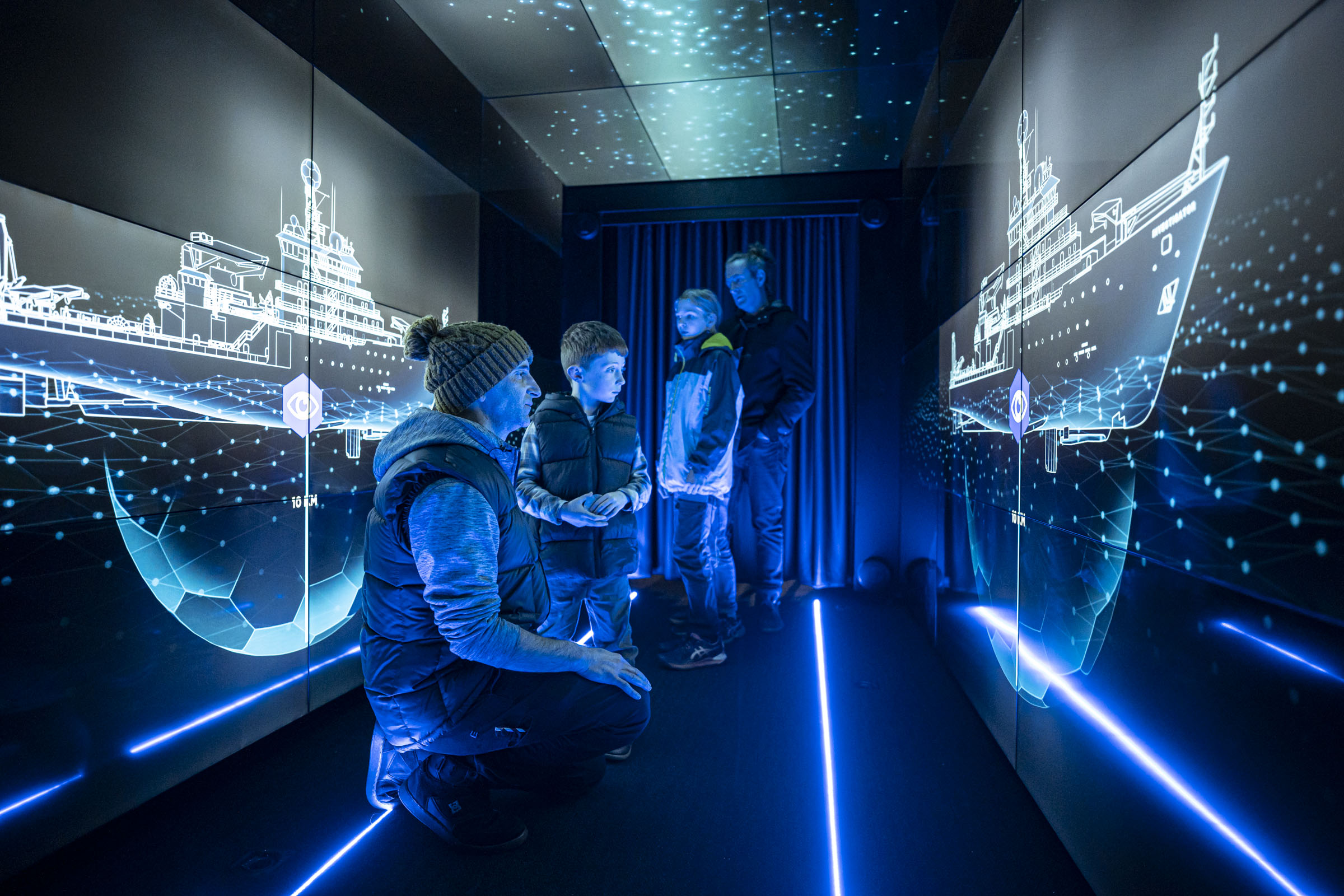 A group of people in a narrow hallway looks at tv screens that surround them.