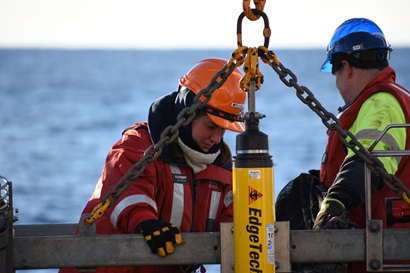 Two people in high vis gear wearing life jackets work on a large piece of scientific equipment with an ocean horizon behind them.