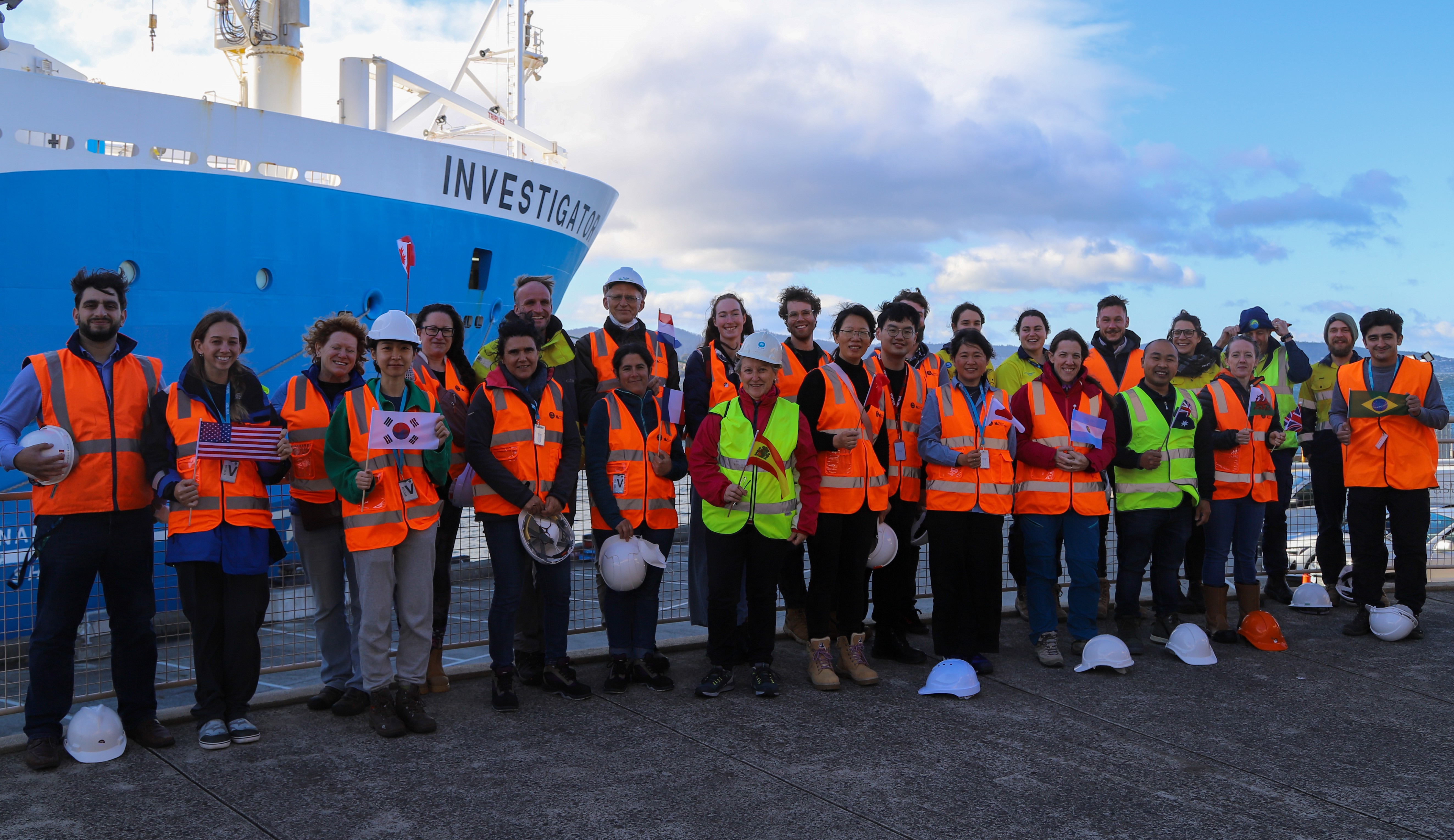 A large group of people in orange and yellow hi-vis vests and some with hard hats stand in front of a ship.