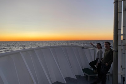 Two people stand on the bow of a ship at sea at sunset.
