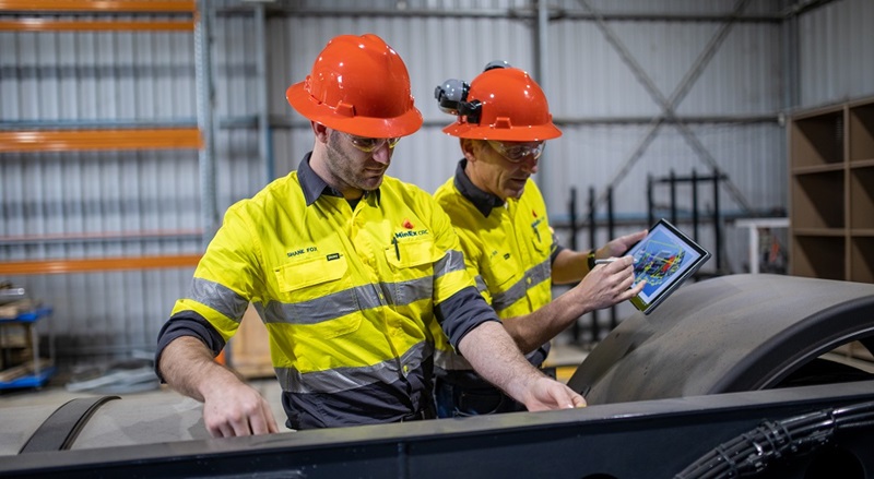 Two men in red hard hats, one measuring a length along machinery and the other holding a tablet device