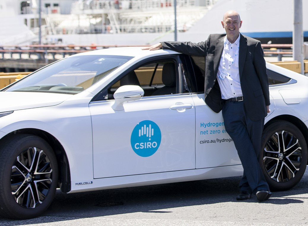 Man in a suit posing in front of a white Toyota Mirai with the CSIRO logo on the door.