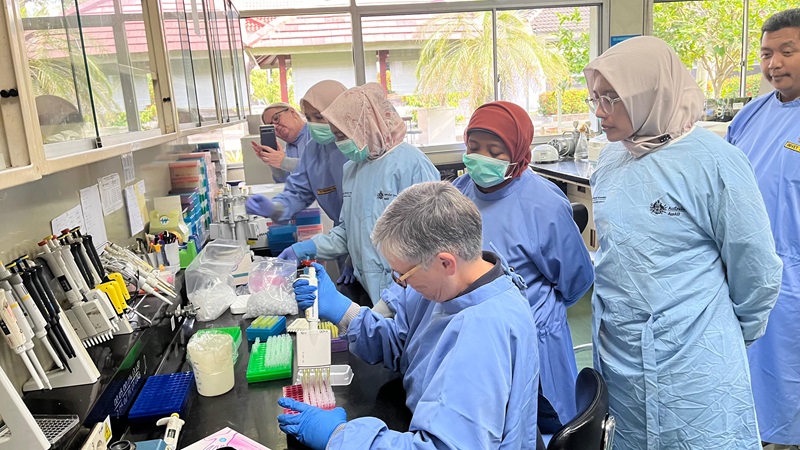 Seven people wearing blue labcoats are either working or observing work in a laboratory.