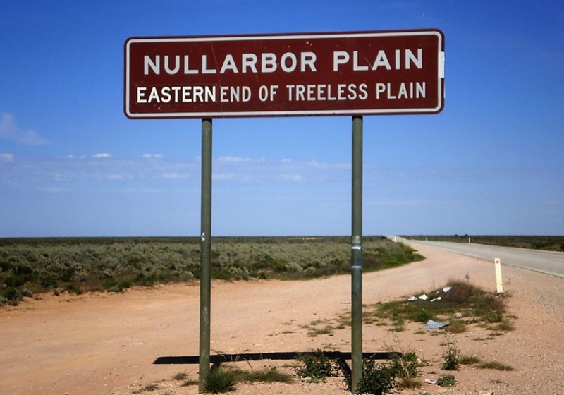 Sign reading Nullarbor plain eastern end of treeless plain, in front of a flat landscape of orange sand and shrubs, under a blue sky