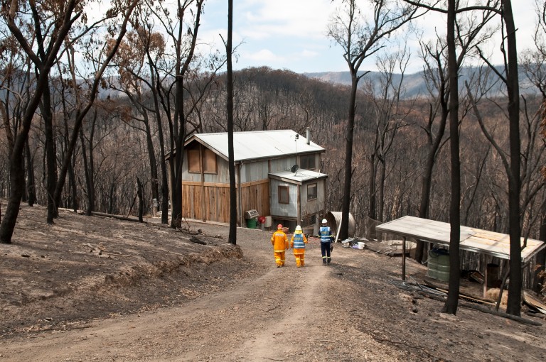 Three firefighters walk down the hill to a house, surrounded by burnt bush.