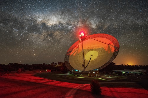 A large radio telescope dish is lit red, with a starry night sky behind