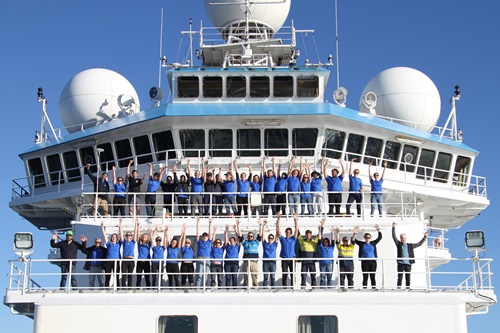 A large group of people in blue t-shirts stand on two decks of a ship at sea.