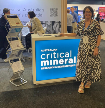 Person standing in front of a backlit cabinet at a conference booth at IMARC. There is a brochure stand to the left and two people in the booth behind talking. Words on the cabinet read 'Australian Critical Minerals Research and Development Hub'. 