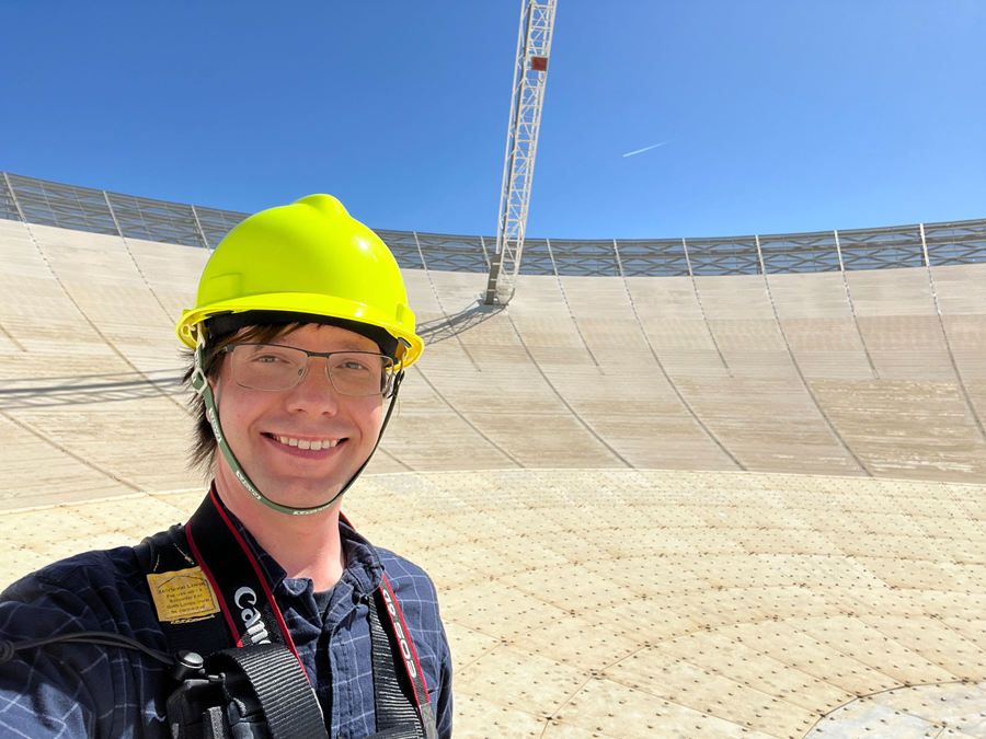 A person in a yellow hard hat smiles at the camera. They are standing on a curved white surface of a radio telescope.