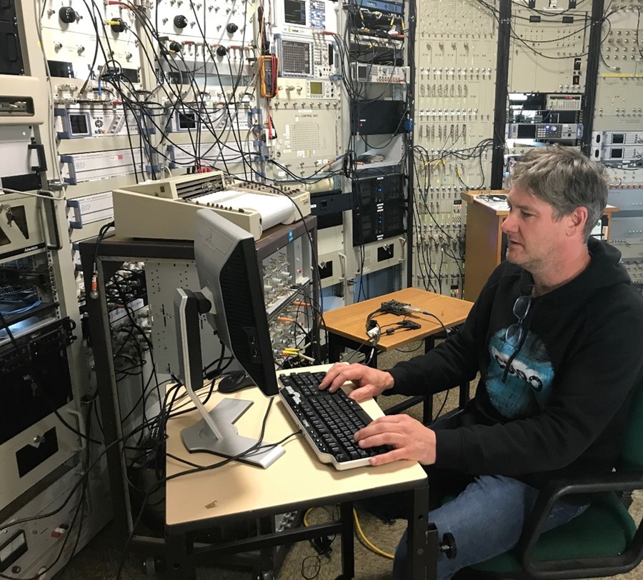 Man sits at a computer with a mess of buttons and cables behind him.