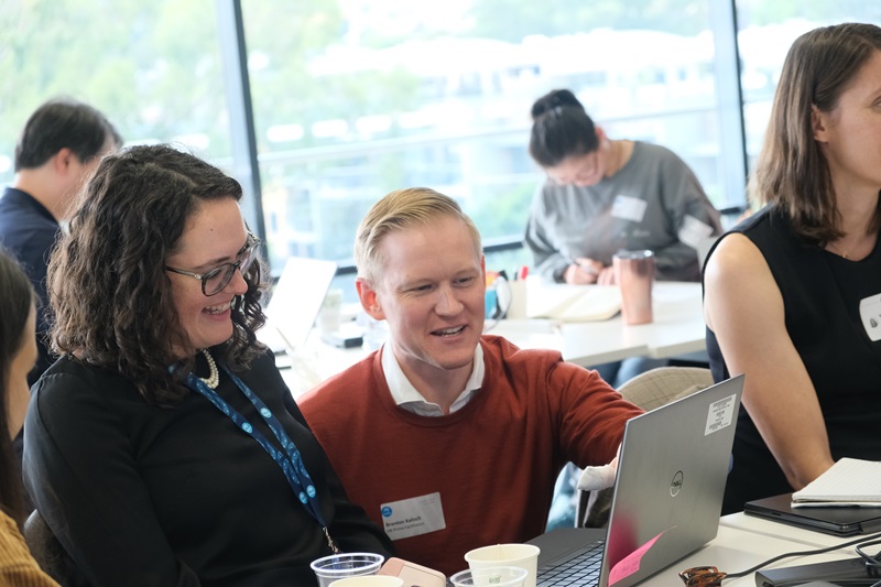 A man and woman are talking while looking at a laptop screen in front of them on the table. 