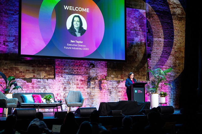 A speaker stands at a podium on a stage with a large screen displaying "Welcome" and "Jen Taylor, Executive Director, Future Industries, CSIRO" against a colorful background.