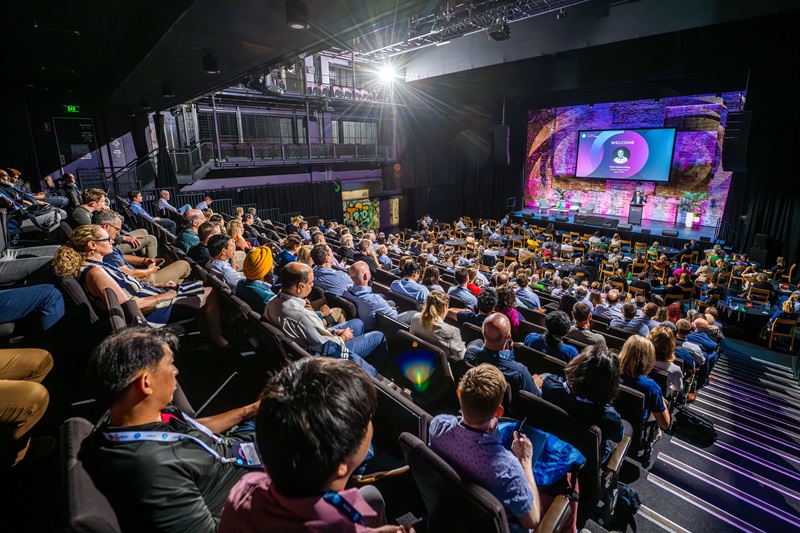 People seated in an auditorium watching a presentation on a stage with a large screen displaying a logo and colorful graphics.