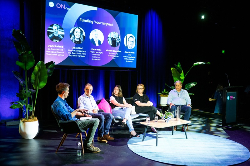 A panel of people sitting on a stage, engaged in discussion, with a screen behind them showcasing relevant content.