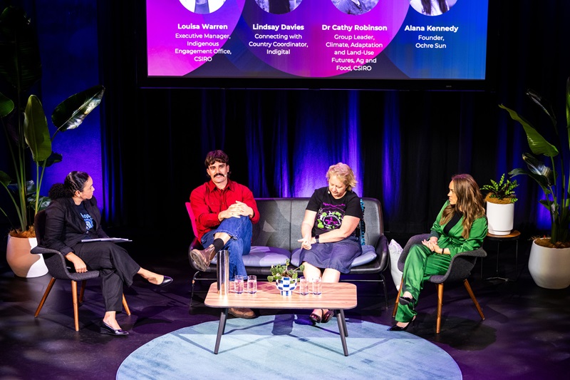  Four participants on a stage, positioned in front of a screen that serves as a backdrop for their presentation.