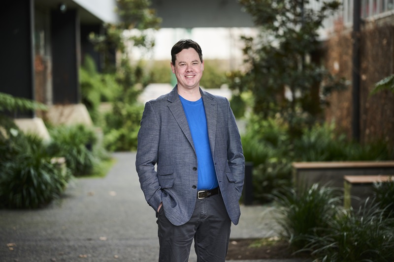 Man in a grey blazer and bright blue shirt stands, hands in pockets and smiling at the camera. Behind him is lots of green plants, potted amongst a brick building 