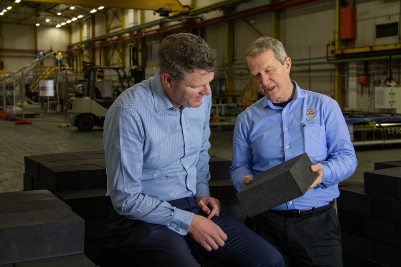 Two men in blue suit shirts are standing together in what might be a warehouse. They look like they are in mid-conversation about the black square object the man on the right is holding. 