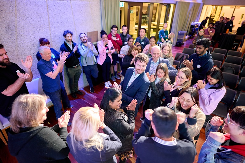 A group of people are standing in an informal circle congratulating and clapping for a couple of excited people in the corner.