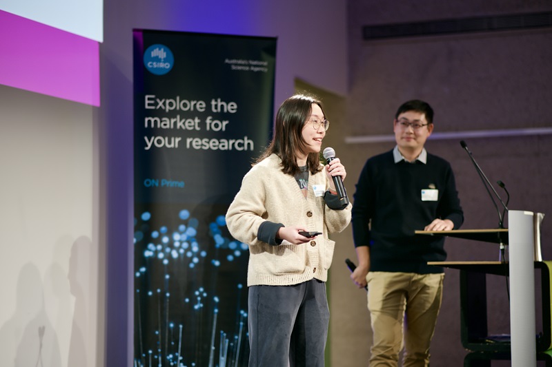 A woman presents an object on stage, with a microphone in one hand and a small remote in another. In the background is a man at the lectern watching her and a standing banner that reads "Explore the market for your research".