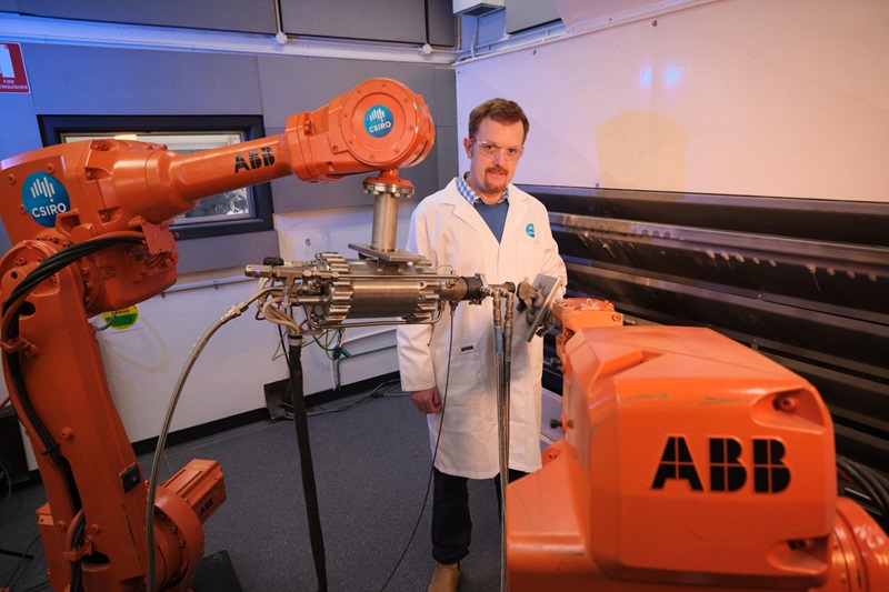 A man in a white CSIRO branded lab coat stands amongst large orange robotic machinery