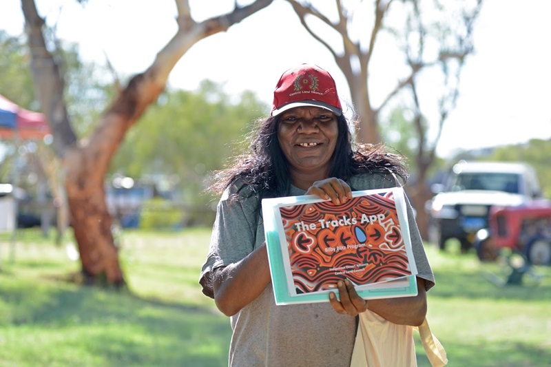 Picture of an Indigenous woman wearing a Central Land Council hat, smiling for the camera and holding up a poster that reads "The Tracks App"