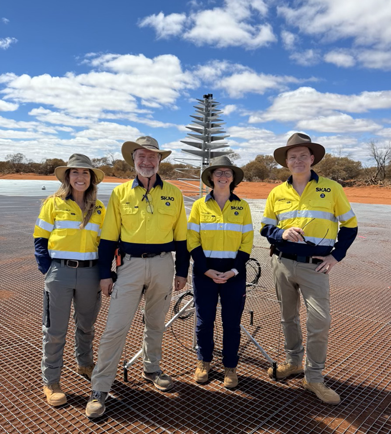 A brunette woman, Angela Teale (far left), stands wearing a yellow high-vis shirt with the SKAO and CSIRO logos on her left chest with three other people in front of the first silver-metal tree-shaped antenna of the SKA-Low telescope on the red soil and steel mesh on Wajarri Country. Credit: SKAO. 